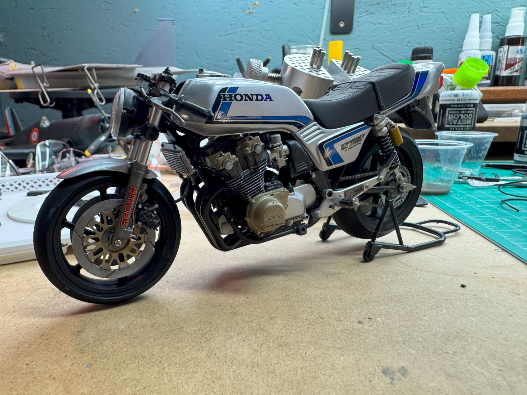 A close-up photo of a detailed scale model motorcycle on a workbench. The model is a Honda, showcasing intricate components such as the wheels, engine, and handlebars, surrounded by various modeling tools and materials.