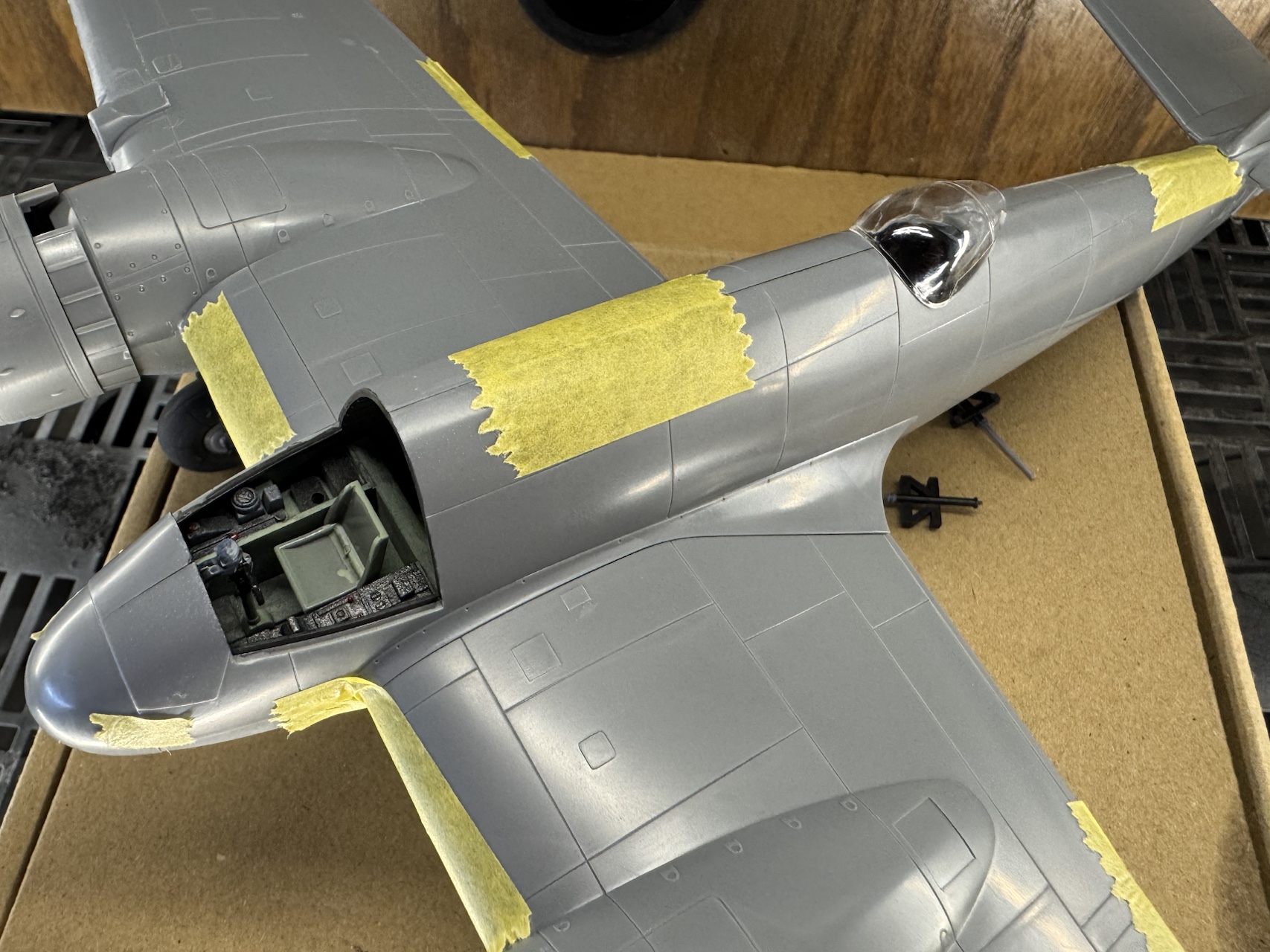 Close-up view of a scale model airplane with masking tape applied on the wings and cockpit area, showing the detail of the unpainted plastic parts.